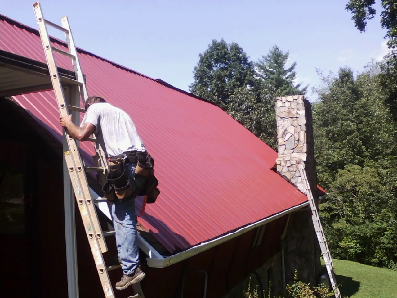 Roofer on a ladder beside a red metal roof and stone chimney — Soffit & Fascia Repair by Roofing Brothers in Duryea
